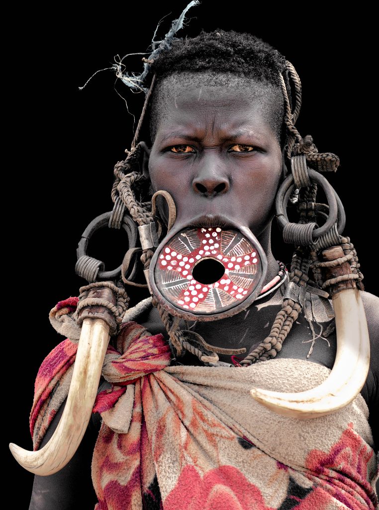 Ein eindringliches Nahaufnahme-Porträt einer Mursi-Frau, die mit einem intensiven, ernsten, ja sogar bösem Blick, direkt in die Kamera schaut. Sie trägt einen großen, traditionellen Lippenteller in ihrer Unterlippe, der mit weißen Punkten und einem roten Ring verziert ist und ein Loch in der Mitte aufweist. Massive Stoßzähne, montiert an schweren Metallringen, hängen von ihren Ohrläppchen herab und rahmen ihr Gesicht ein, während sie auf ihren Schultern ruhen