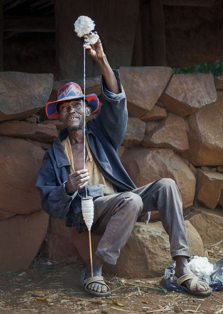 Ein lebendiges Farbfoto eines älteren Konso-Mannes aus Äthiopien bei der traditionellen Handarbeit. Er trägt den charakteristischen, eng anliegenden Hut der Konso, der mit einem markanten grafischen Muster in Blau und Rot verziert ist. In seiner einen Hand hält er eine hölzerne Spindel, während die andere Hand den Baumwollfaden weit nach oben ausgestreckt hält, um die Spannung beim Spinnen zu kontrollieren. Sein Gesicht ist konzentriert und von der Sonne gegerbt. Er sitzt vor einer traditionellen Steinmauer. Das warme Tageslicht betont die Details seiner arbeitenden Hände und die feine Struktur des Fadens.
