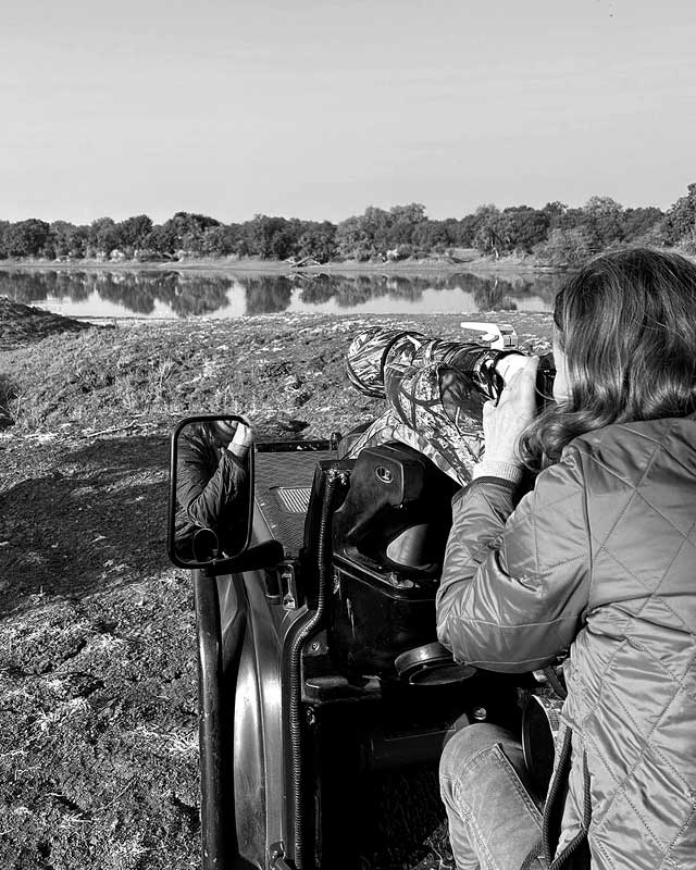 Schwarz-Weiß-Foto von Barbara Westphalen, Ansicht von Hinten, die durch ihr Teleobjektiv aus einem Jeep auf ein Wasserloch schaut und fokusiert.
