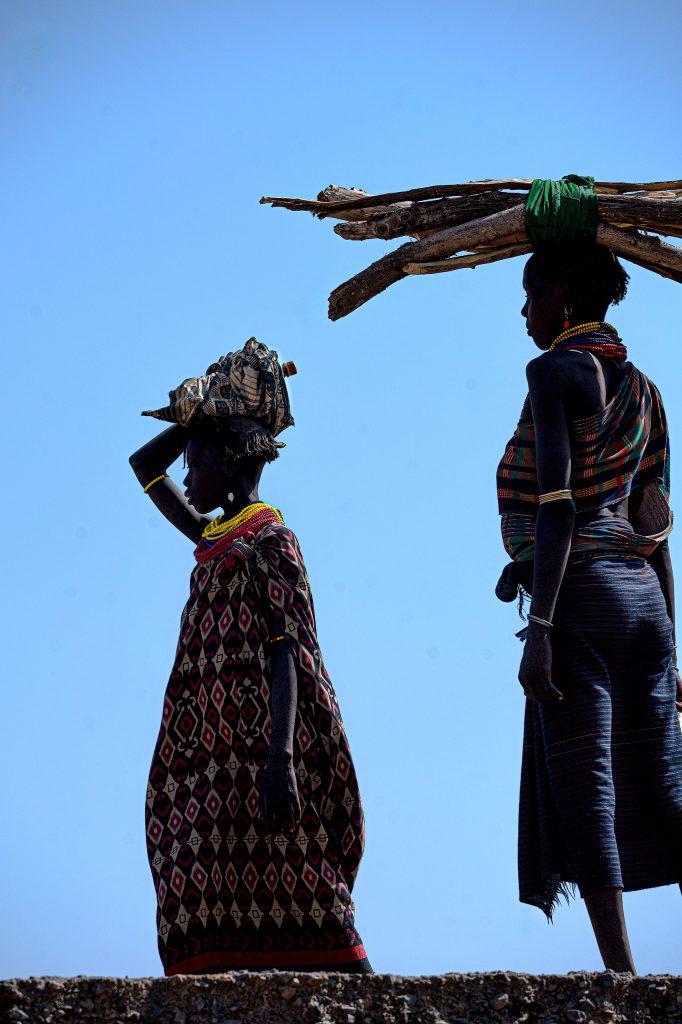 Silhouetten von zwei afrikanischen Frauen, von unten gegen einen blauen Himmel fotografiert, die schwere Holzbündel auf ihren Köpfen tragen; in den dunklen Schatten sind noch Details ihrer farbenfrohen Kleidung zu erkennen.