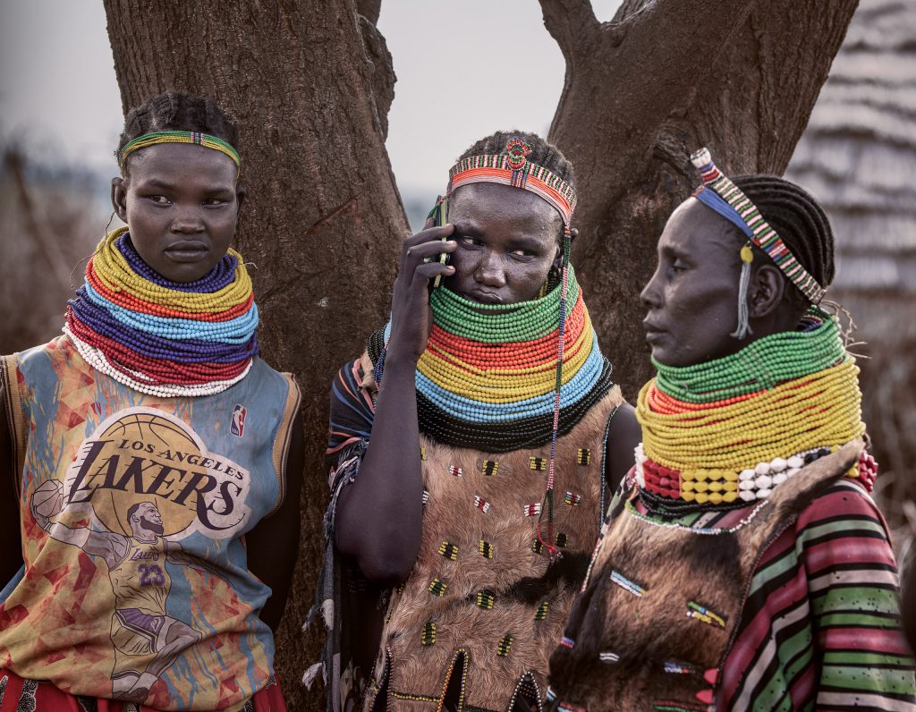 Spannender Kontrast aus Tradition und Moderne: Drei junge Nyangatom-Frauen mit schwerem, traditionellem Halsschmuck, während eine ein Smartphone benutzt.