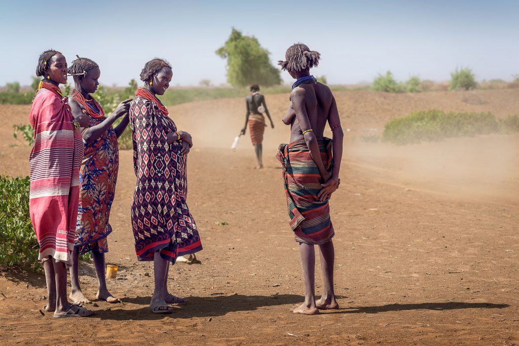 Frauen vom Stamm der Dassanech in traditionellen Tüchern stehen in der Savanne und blicken einer weiteren Frau hinterher, die einen staubigen Weg entlang in die Ferne läuft.