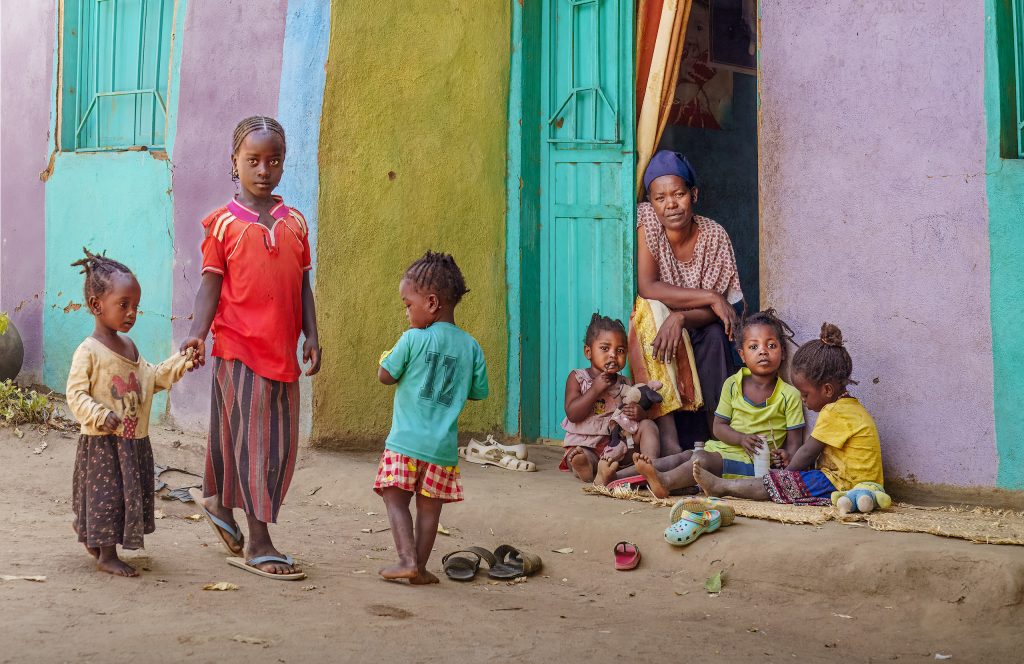 Fine Art Travel Fotografie von einer sehr farbenfrohen Szene vor einer bunten Hausfassade: Eine ältere Frau vom Stamm der Ari sitzt im Hauseingang, während davor viele Kinder miteinander spielen.