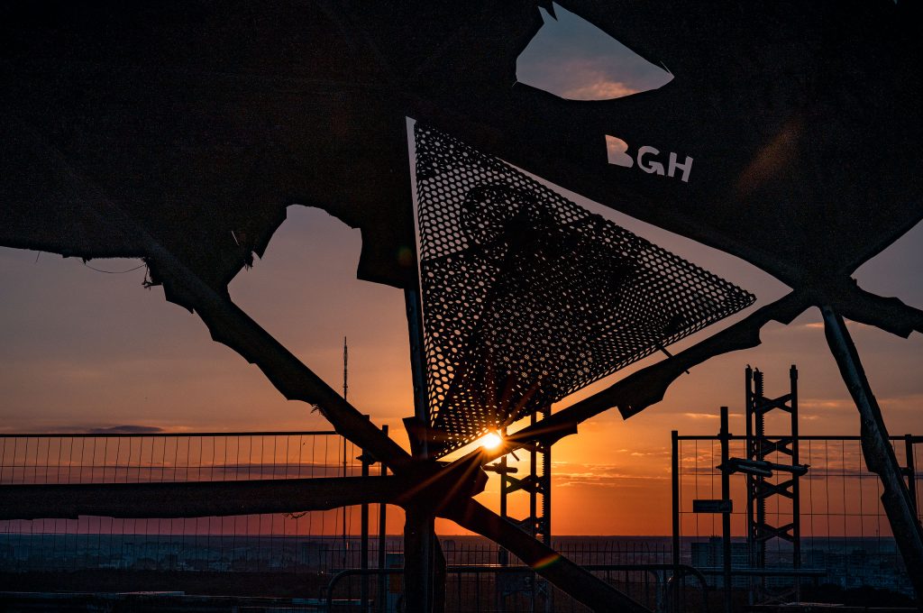 Fine-Art-Fotografie: Abstraktes, schwarzes Blechelement mit der eingestanzten Aufschrift „BGH“ als Silhouette vor einem farbenfrohen Sonnenuntergang.