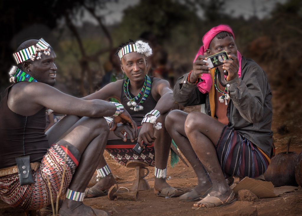 Fine-Art-Travel-Fotografie: Drei junge Männer vom Stamm der Hamer mit dem traditionellen Perlenschmuck der Junggesellen sitzen auf ihren hölzernen Kopfstützen, während einer von ihnen mit dem Handy fotografiert.