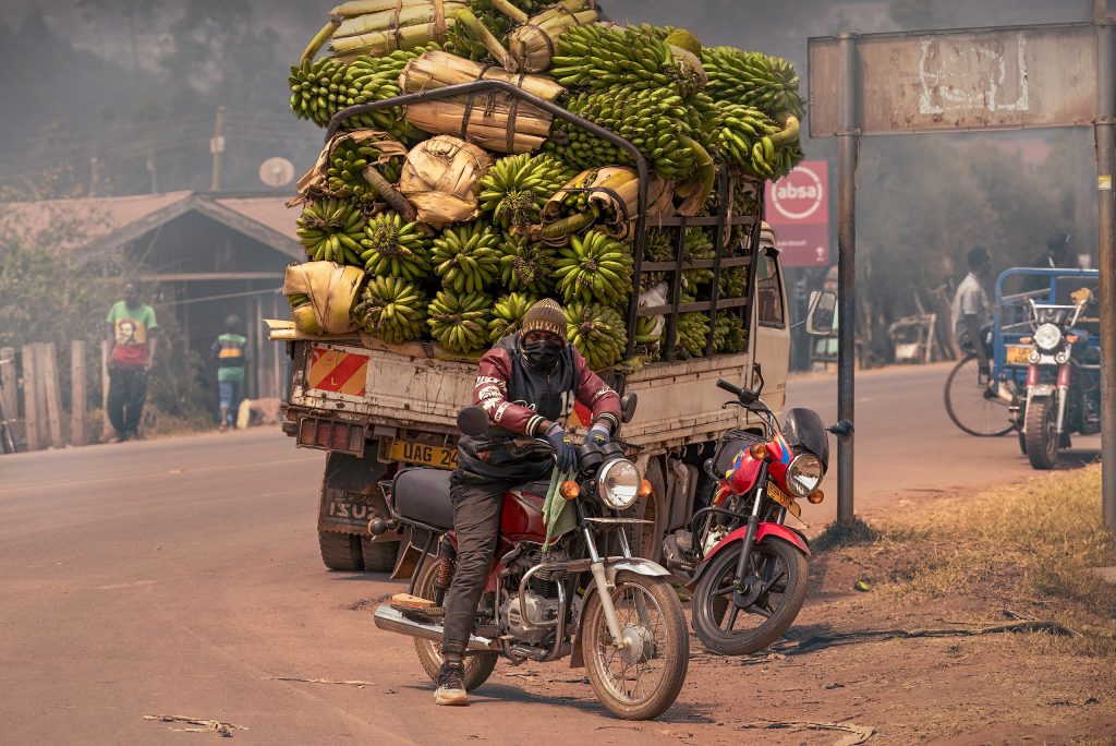 Straßenszene in Uganda: Ein Mann auf einem Motorrad fährt vor einem Lastwagen her, der mit Unmengen von Bananen überladen ist; der Hintergrund ist dunstig und staubig.