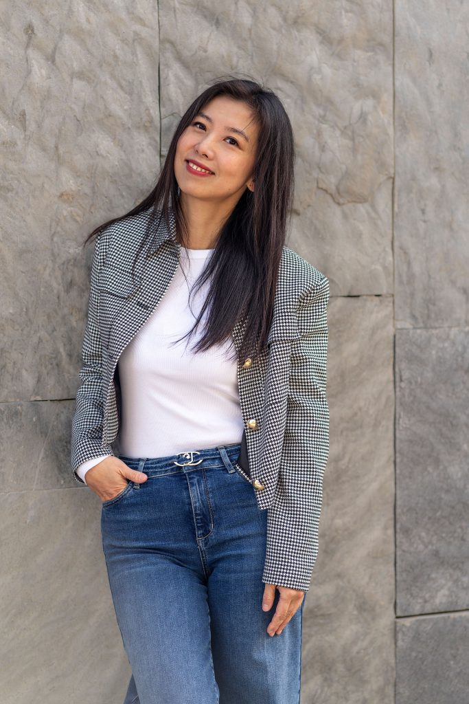 Professionelles Porträt einer asiatischen Frau mit langen schwarzen Haaren. Sie lächelt freundlich in die Kamera und trägt eine klassisch karierte Blazer-Jacke vor einem neutralen Hintergrund.