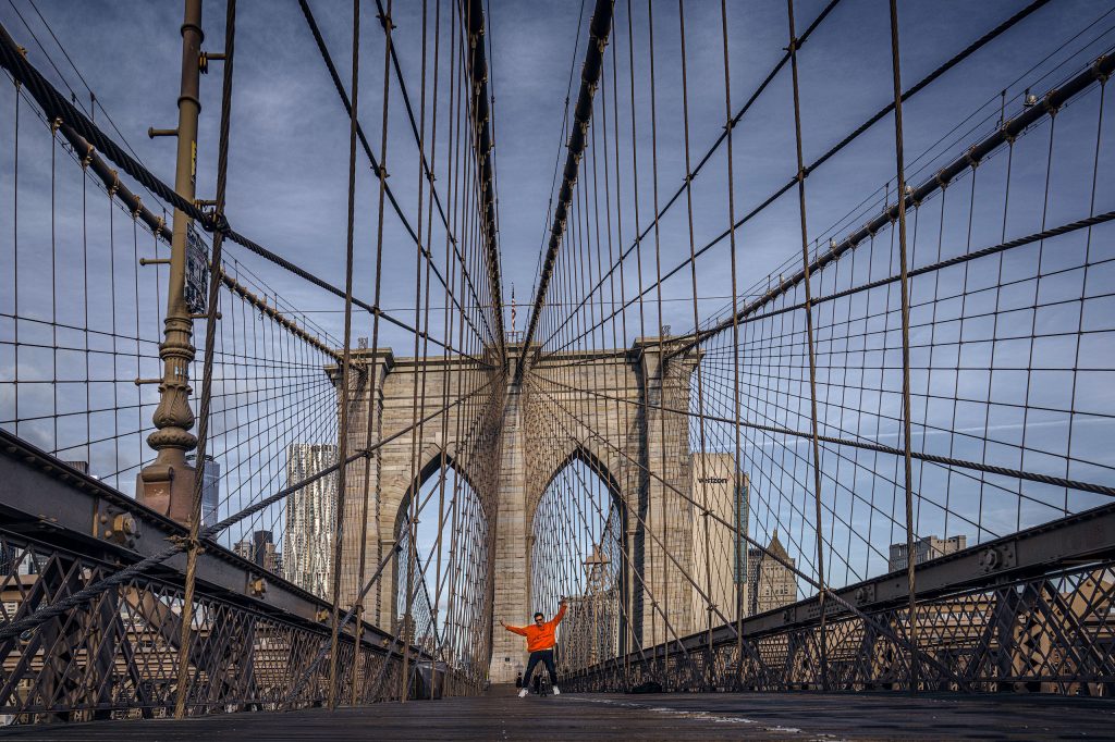 Fine-Art-Stadtfotografie: Ein junger Mann in einem orangenen Hoodie tanzt auf dem Fußgängerweg in der Mitte der Brooklyn Bridge, umgeben von der ikonischen Seilkonstruktion.