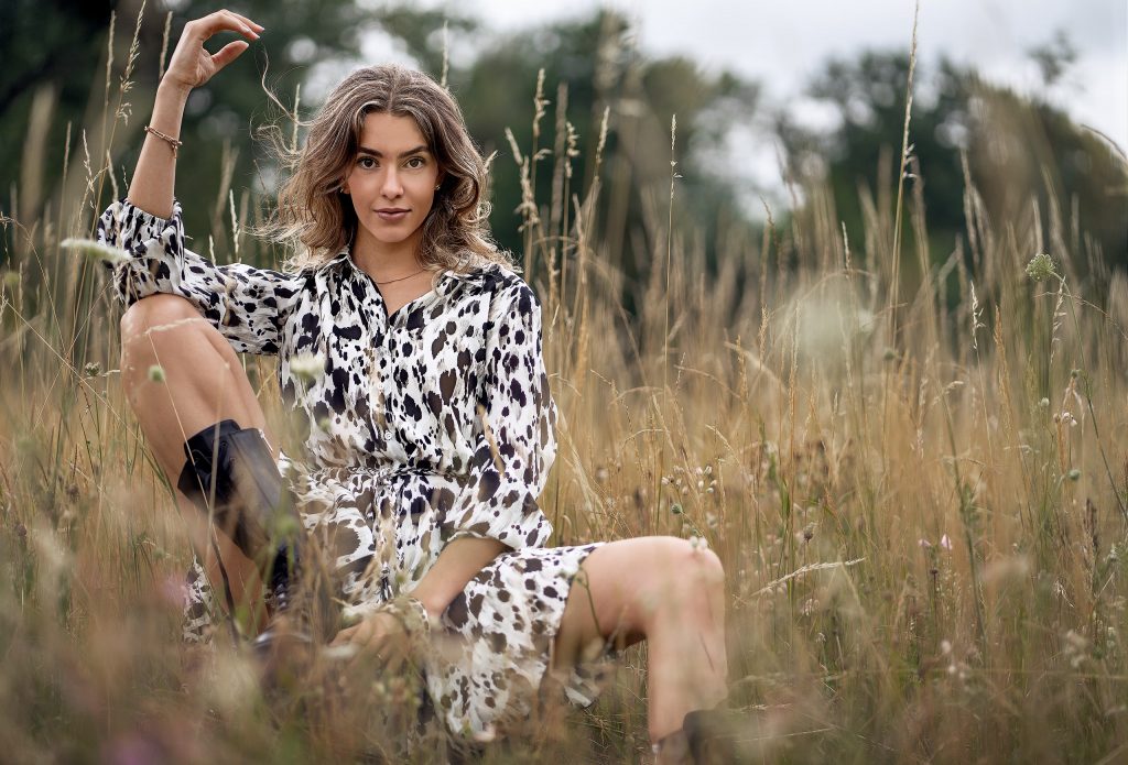 Verträumtes Outdoor-Modefoto, aufgenommen durch unscharfe, hohe Halme einer spätsommerlichen, gelblichen Wiese. Im Fokus steht ein Modell in einem Sommerkleid mit markantem, schwarzem Fleckendruck. Dazu trägt sie als derben Stilbruch dunkle, klobige Chunky Boots. Die verschwommene Perspektive im Vordergrund verleiht dem Bild Tiefe und eine geheimnisvolle Stimmung.