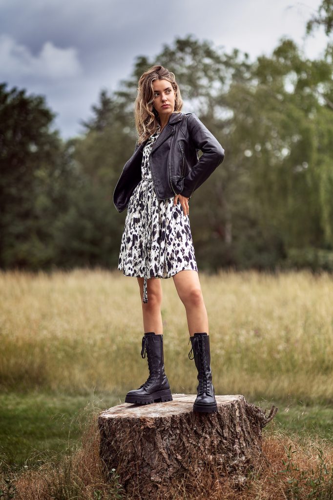 Mode-Shooting auf einer Wiese: Modell in einem Sommerkleid mit Animal-Print und einer schwarzen Biker-Lederjacke. Sie steht auf einem Baumstumpf und blickt herausfordernd zur Seite. Fokus auf den Stilbruch der Outfits. Die natürliche Umgebung kontrastiert stark mit dem urbanen Look.