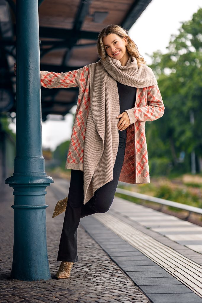 Mode-Shooting am Bahnhof: Blondes Modell mit langen Locken posiert an einer Säule auf dem Bahnsteig. Sie trägt einen karierten Strickmantel und einen groben Strickschal vor einem natürlichen Hintergrund mit Bäumen.