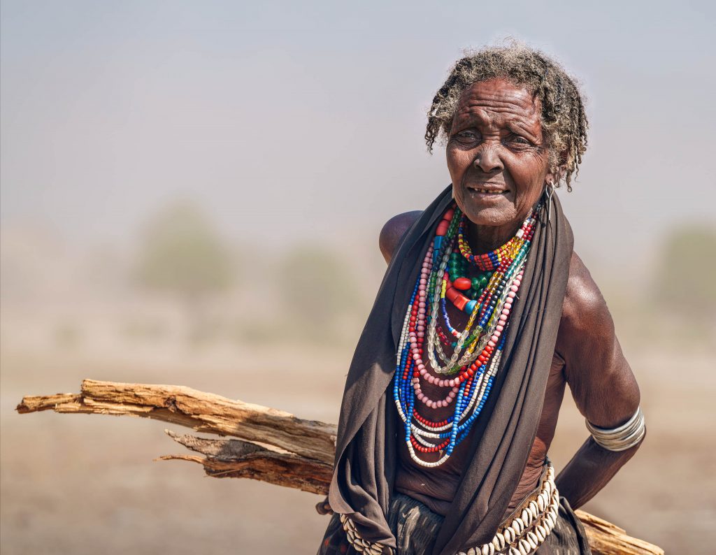 Charakterportrait einer älteren Arbore-Frau in traditionellem Halsschmuck; das faltige Gesicht und das Stück Holz in ihrer Hand betonen die Struktur und Tiefe der Fine-Art-Fotografie.