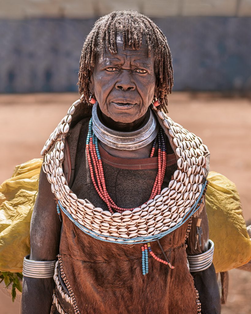 Fine-Art-Portrait einer älteren Frau vom Stamm der Hamer; sie trägt den traditionellen Schmuck verheirateter Frauen, bestehend aus einem breiten Ledergürtel mit Kaurischnecken und den typischen schweren Metallreifen um den Hals.