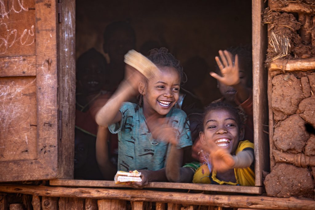 Fine-Art-Fotografie einer fröhlichen Kinderszene in einer äthiopischen Schule: Eine Gruppe strahlender Kinder schaut aus dem Fenster eines traditionellen Lehmgebäudes und winkt fröhlich in die Kamera.