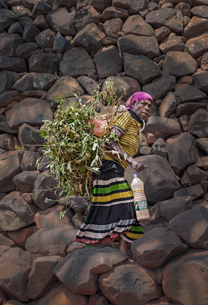 Fine-Art-Fotografie einer Frau in einer farbenfrohen, traditionellen Tracht, die seitlich an einer Mauer entlang schmale Stufen nach oben geht und dabei schwer mit Ästen und Holz beladen ist.