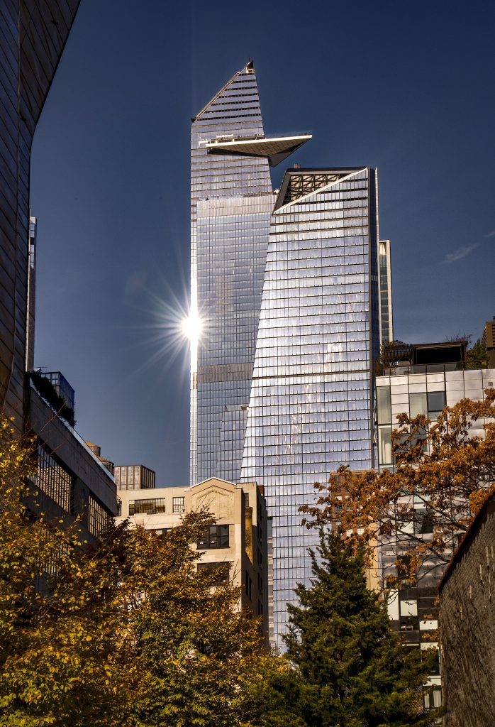Architekturfoto im Licht der Mittagssonne, aufgenommen von der High Line .Ein moderner Wolkenkratzer ragt steil in den tiefblauen Himmel. Das grelle Sonnenlicht bricht sich millimetergenau an der scharfen Glaskante des Gebäudes und erzeugt einen brillanten, strahlenförmigen Sonnenstern Das Zusammenspiel aus geometrischer Härte und dem funkelnden Lichteffekt verleiht der urbanen Szene eine dynamische, fast futuristische Energie.