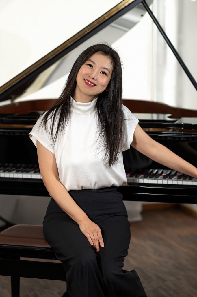 Asiatische Pianistin im weißen Seidentop sitzend vor einem Flügel in einem hellen Raum mit Fenster.