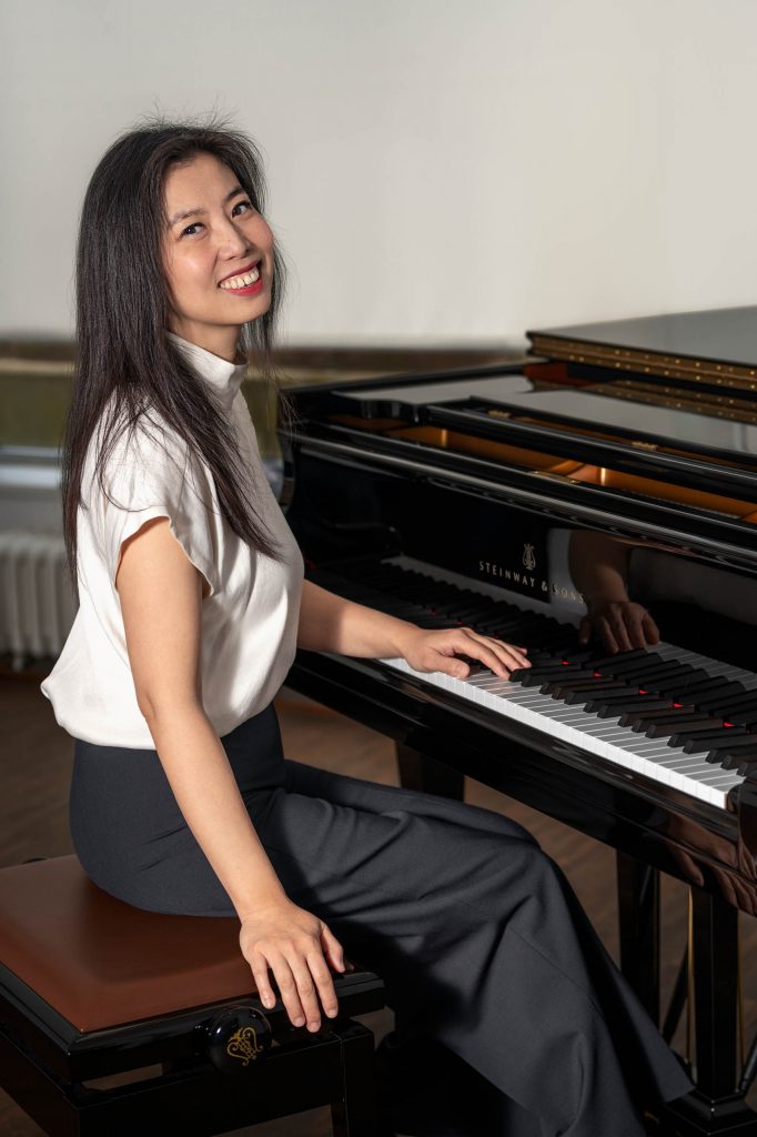Porträt einer asiatischen Pianistin im weißen Seidentop. Sie sitzt vor einem Flügel in einem hellen Zimmer und blickt lächelnd in die Kamera, eine Hand ruht auf den Klavier Tasten.