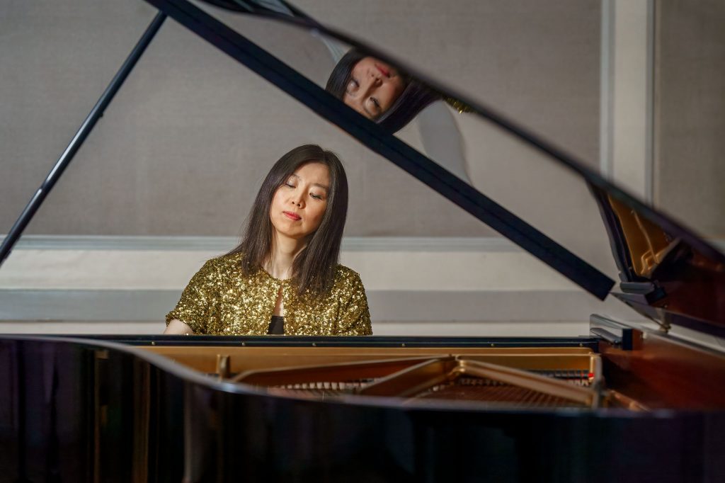 Eine asiatische Pianistin in einer auffälligen, goldenen Paillettenjacke spielt Klavier. Die Aufnahme erfolgt kreativ durch den geöffneten Flügeldeckel, auf dem sich ihr Gesicht spiegelt.