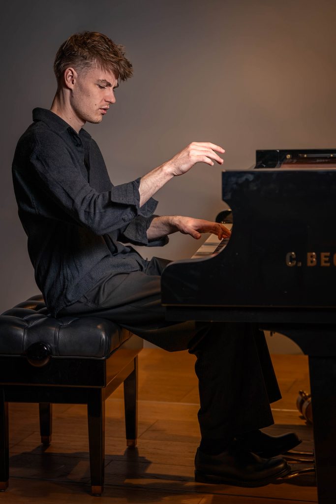 Seitliche Aufnahme eines Pianisten in schwarzer Kleidung, der versunken am Klavier in einem dunklen Raum spielt.