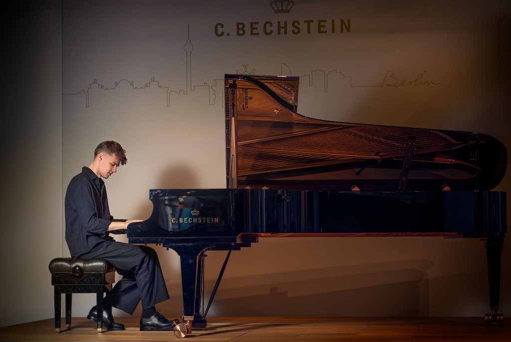 Atmosphärisches Künstlerporträt von Pianisten Janik Chêch in einer dunklen Kulisse. Er spielt am Flügel positioniert, der durch gezielte Beleuchtung hell erstrahlt und im starken Kontrast zur tiefen, schattigen Umgebung steht. Die geheimnisvolle und fokussierte Lichtstimmung betont die Verbindung zwischen Musiker und Instrument.