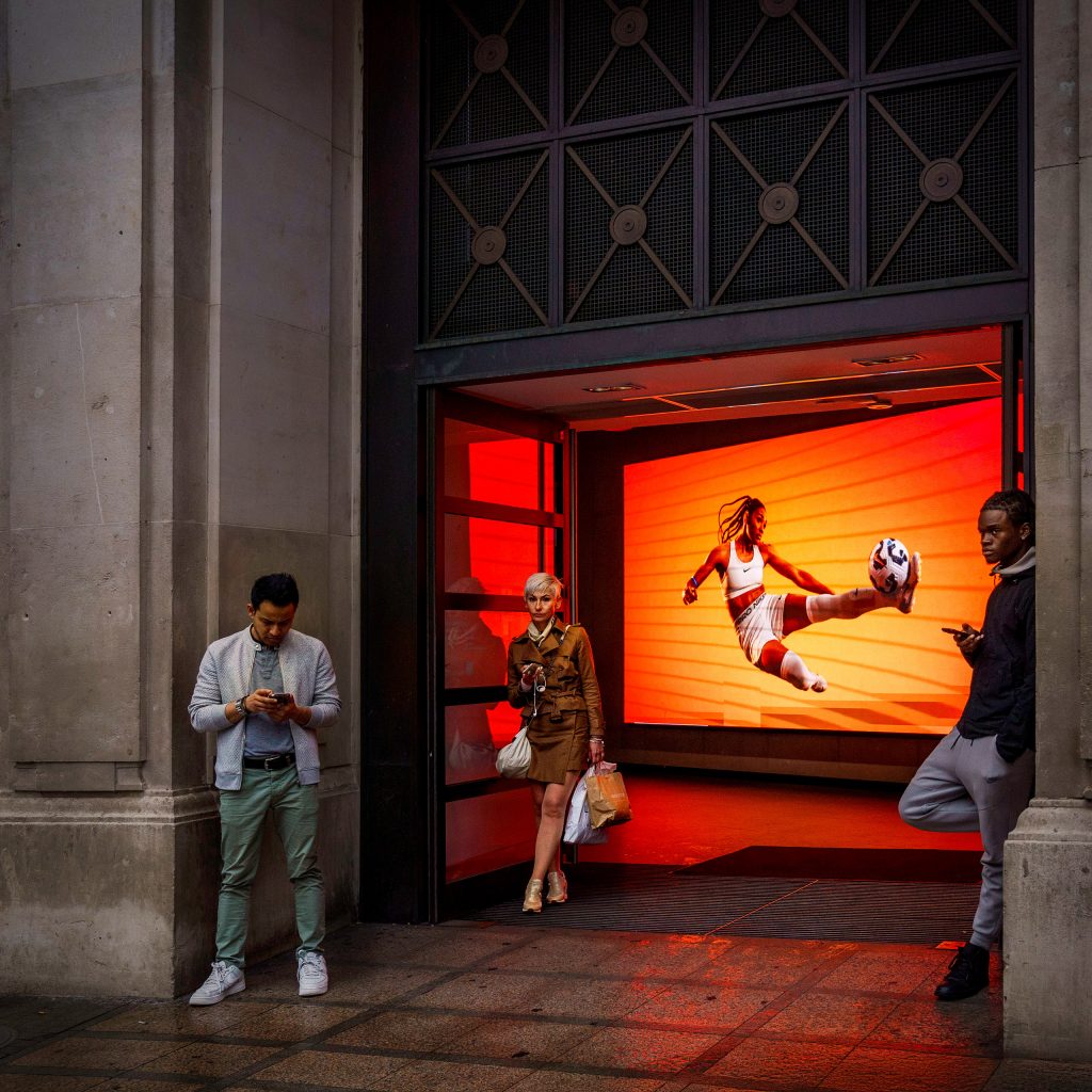 Streetphotography London: Eine Frau mit Einkaufstüte geht vor einem leuchtend orangen Bildschirm, der einen Fußballspieler in dynamischer Pose zeigt, aus dem Laden. Ein Mann links und ein Mann rechts stehen daneben und schauen auf ihre Smartphones.