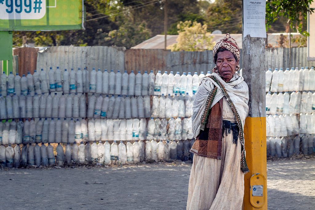 Fine-Art-Fotografie einer Straßenszene in Afrika: Eine ältere Frau lehnt nachdenklich an einem Holzpfahl, während im Hintergrund ein Wellblechdach und ein kunstvoller Zaun aus alten Plastikflaschen zu sehen sind.