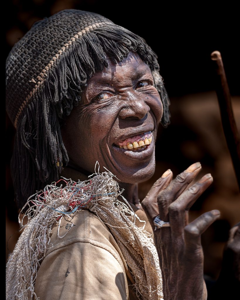 Ausdrucksstarkes, dunkles Fine-Art-Foto einer älteren Konso-Frau, die herzlich lacht; ihr Blick ist seitlich zur Kamera gerichtet und auf dem Kopf trägt sie eine auffällige, gehäkelte Mütze mit Fransen.