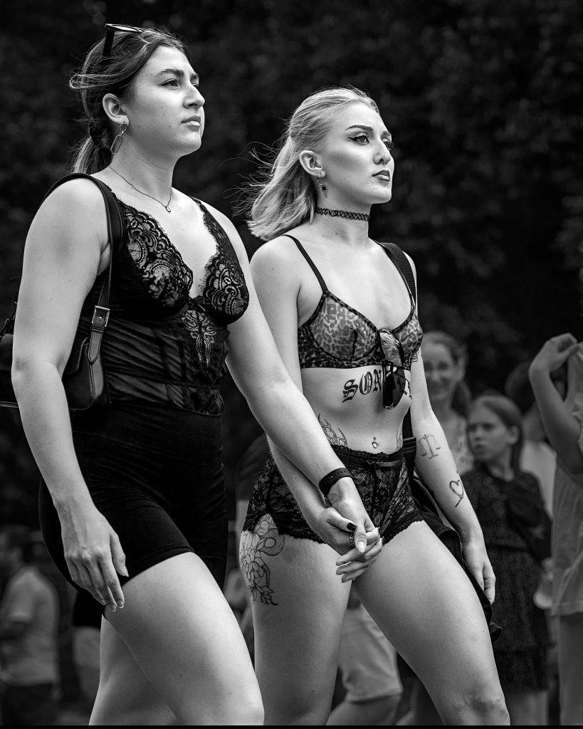 Ausdrucksstarke Schwarz-Weiß-Streetphotography auf der Loveparade. Zwei junge Frauen mit markanten Tätowierungen gehen Hand in Hand durch eine belebte Straße. Sie tragen aufreizende, schwarze Reizwäsche und blicken mit einem selbstbewussten, herausfordernden und direkten Ausdruckvor sich hin. Die dynamische Bewegung und die kontrastreiche Ästhetik fangen die freie, energiegeladene Atmosphäre des Straßenumzugs perfekt ein.