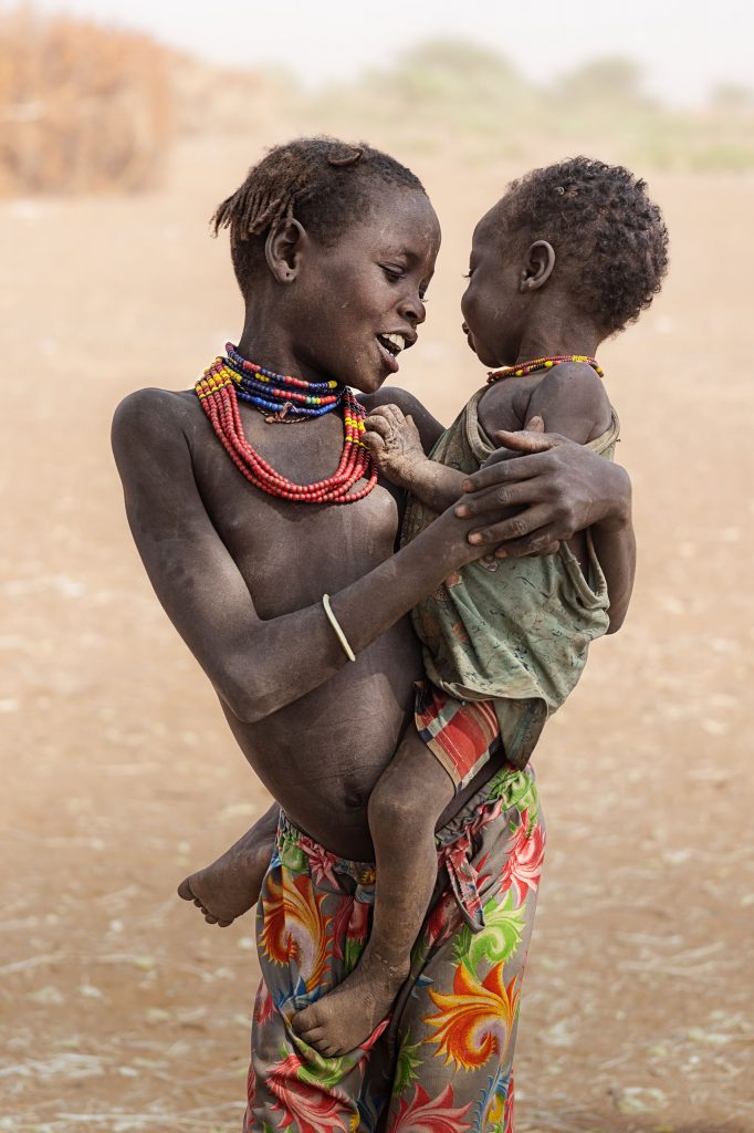 Inniger Moment in der Savanne: Ein älteres afrikanisches Mädchen trägt ihr kleines Geschwisterchen auf dem Arm und schenkt ihm ein zärtliches, fröhliches Lachen; der Hintergrund ist in ein staubiges Gelb getaucht.s