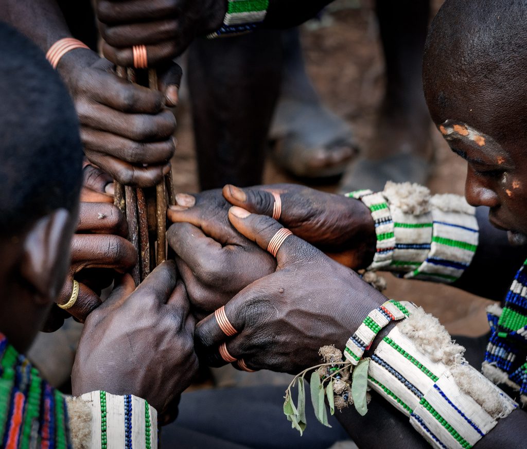 Detailaufnahme Fine Art Foto. Ein kraftvoller Moment der Gemeinschaft bei einer afrikanischen Bullensprung-Zeremonie, bei dem mit traditionellen Perlenbändern geschmückte Männerhände gemeinsam einen Zeremonie Stab halten.