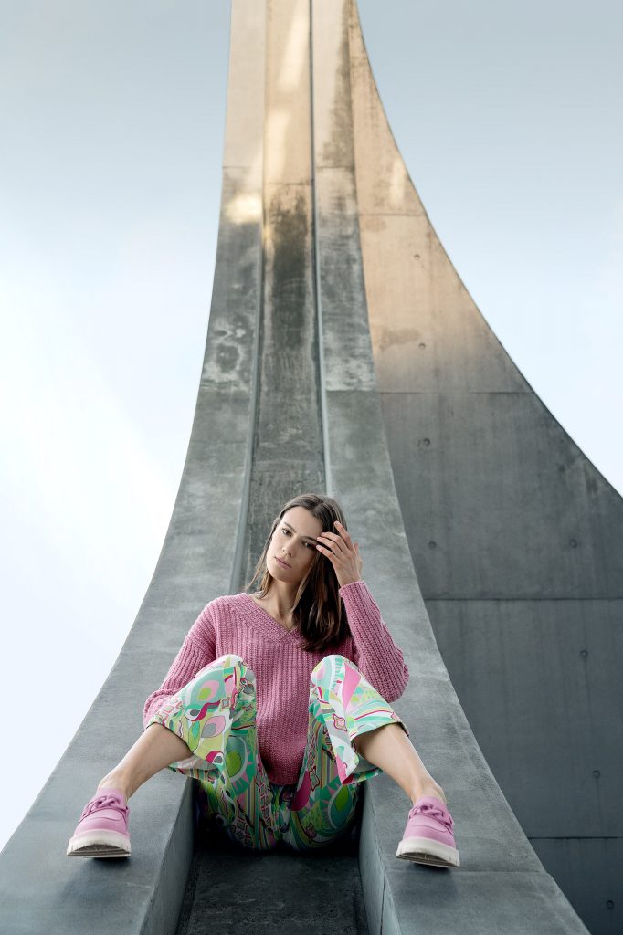 Minimalistisches Fashion-Porträt vor einer Kulisse moderner Architektur mit klaren Linien und Betonstrukturen. Ein Modell sitzt entspannt auf einem architektonischen Element und trägt einen auffälligen, groben pinken Pullover im Kontrast zu einer Hose mit markantem, grafischem Muster.