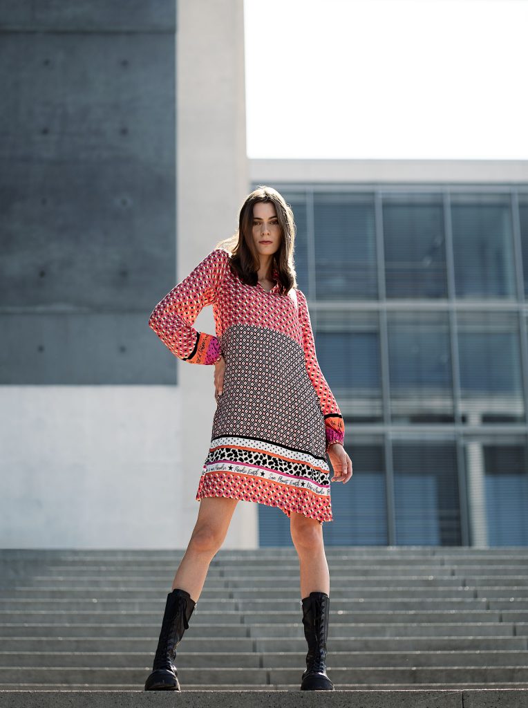 Urbanes Mode-Shooting vor moderner Architektur: Modell in einem Kleid mit grafischem Muster und derben, schwarzen Schnürboots. Stilvoller Kontrast aus femininem Design und robusten Boots.
