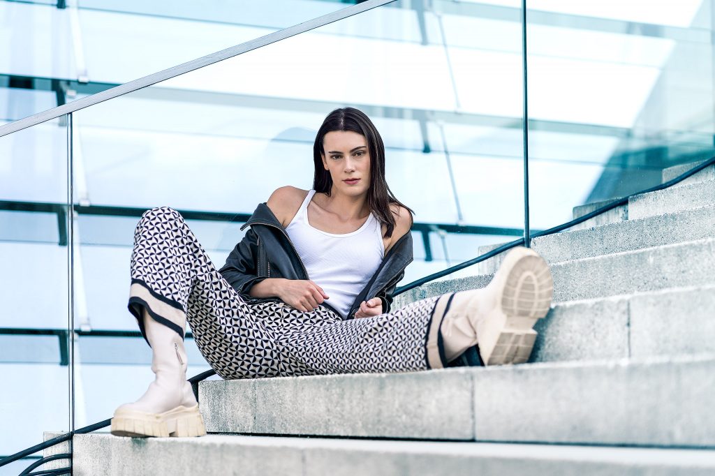 Mode-Shooting vor moderner Glasfassade im kühlen Farbton. Dunkelhaariges Model posiert provokant auf einer Treppe in einer Kombination aus schwarzer Lederjacke, Hose mit grafischem Muster, weißem Top und derben Boots.