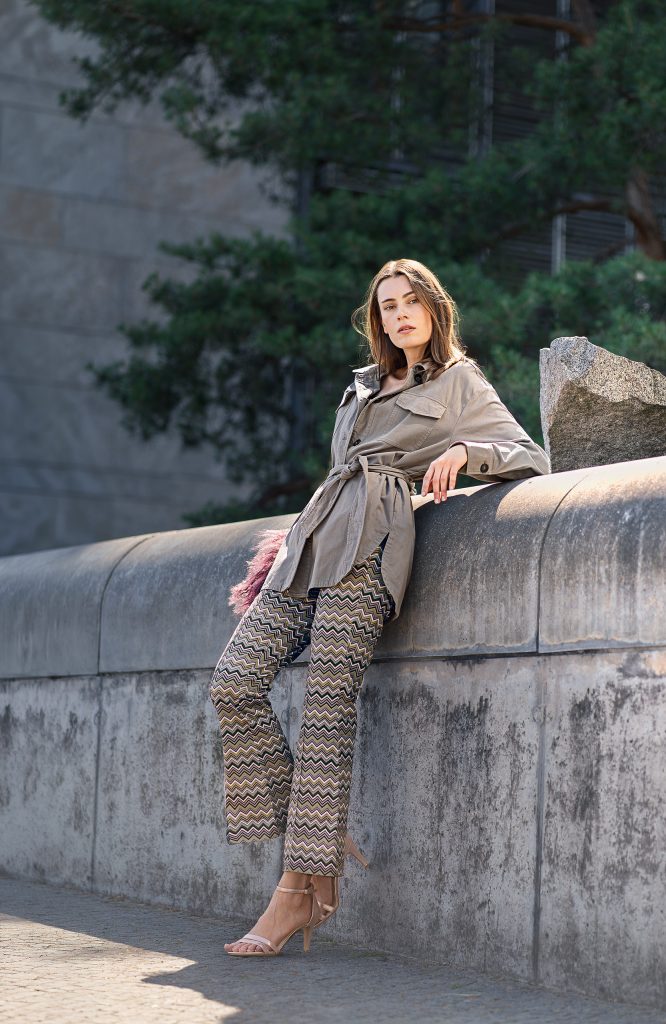 Urbanes Mode-Shooting: Modell in einer auffälligen Hose mit grafischem Muster und sportlicher Jacke lehnt lässig an einer Betonmauer vor städtischer Kulisse.