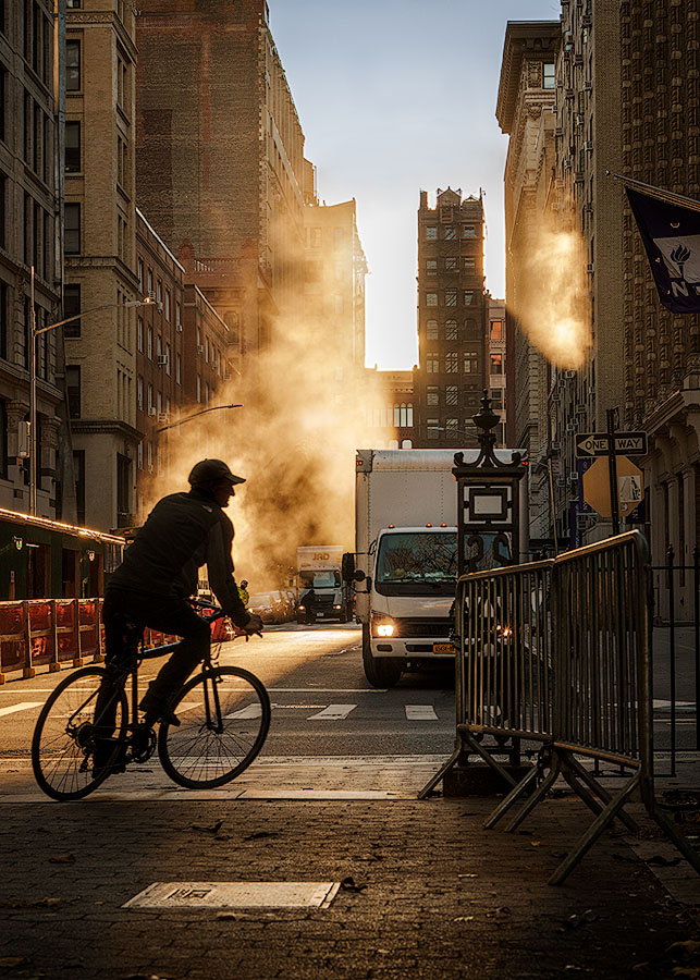 Streetphotography aus dem New Yorker Stadtteil Soho im frühen Morgenlicht. Aus einem Hydranten auf der linken Straßenseite steigt eine dichte, helle Dampfwolke empor, die vom Gegenlicht der tiefstehenden Sonne intensiv erleuchtet wird. Im Vordergrund fährt ein Radfahrer dynamisch als dunkle, scharf gezeichnete Silhouette durch das Bild. Die Pflastersteine der Straße glänzen im Licht. Im Hintergrund ragen die typischen, historischen Soho-Gebäude empor, während die Straße im Dunst des Morgens verschwindet.