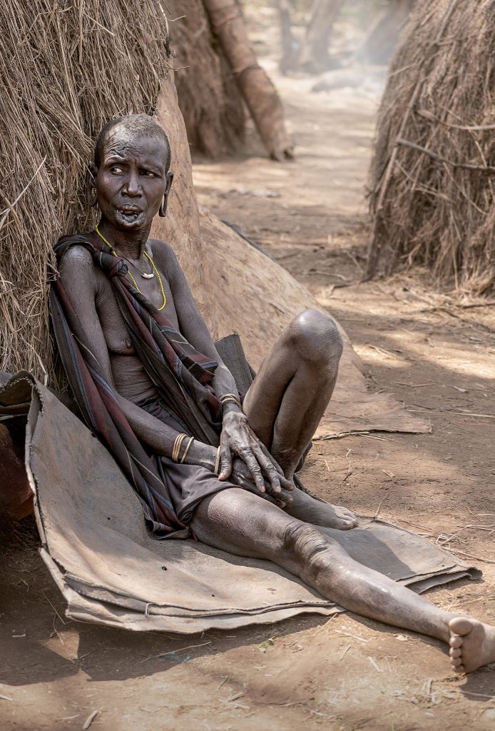 Fine-Art-Portrait einer älteren Mursi-Frau, die mit resigniertem Ausdruck auf dem staubigen Boden sitzt, den Rücken an eine einfache Strohhütte gelehnt; ihre Unterlippe ist traditionell aufgeschlitzt, trägt jedoch keinen Lippenteller.