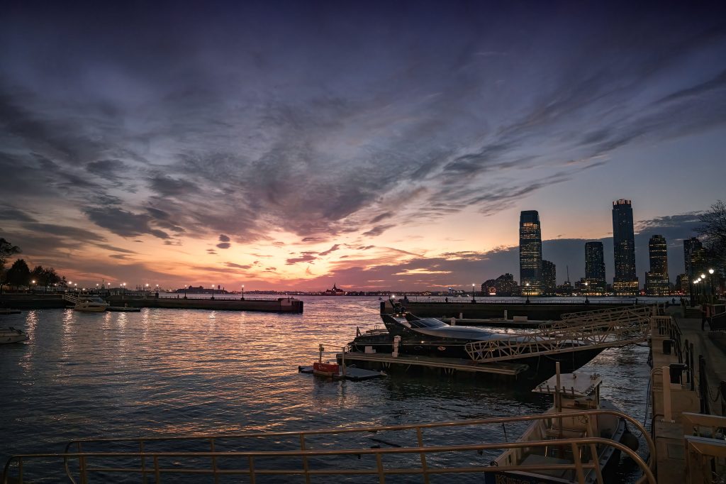 Malerische Fine-Art-Fotografie des North Cove Yacht Harbor in Lower Manhattan bei Sonnenuntergang. Im ruhigen, spiegelnden Wasser des Hafens liegen einige Boote friedlich vor Anker. Der weite Himmel darüber glüht in einem intensiven Farbverlauf aus warmem Orange, sanftem Rosa und tiefem Violett. Am Horizont zeichnen sich die erleuchteten Silhouetten der Hochhäuser des gegenüber liegenden Ufers ab.