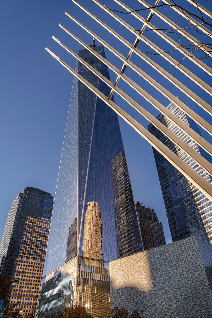 Dynamische Fine-Art-Architekturfotografie in lebendigen Farben am One World Trade Center in New York. Die Perspektive ist nach oben gerichtet und zeigt die Glasfassade eines modernen Wolkenkratzers, in der sich der andere Gebäude spiegeln. Von der Seite ragt ein strahlend weißer, geschwungener Flügel der markanten Oculus-Konstruktion elegant in das Bild hinein. Das Zusammenspiel aus den strengen, reflektierenden Linien des Hochhauses und der organisch wirkenden, weißen Struktur des Flügels erzeugt eine spannungsreiche, zukunftsorientierte Komposition.