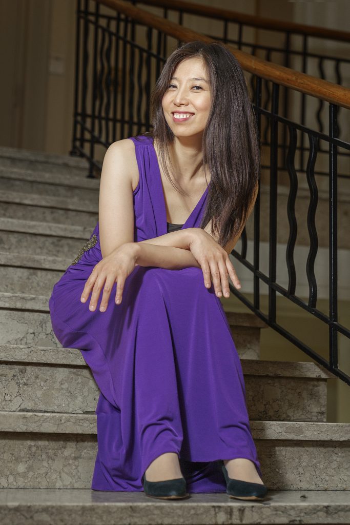 Image-Porträt der Pianistin Huijing Han. Sie sitzt entspannt auf einer Treppe, trägt ein festliches, lila Abendkleid und blickt mit einem herzlichen, natürlichen Lächeln direkt in die Kamera. Die Kombination aus dem glamourösen Kleid und der ungezwungenen Pose auf den Stufen verleiht dem Bild eine moderne Dynamik.