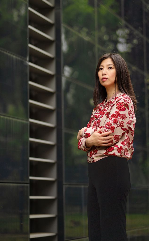Selbstbewusstes Fine-Art-Porträt einer jungen asiatischen Frau. Ein stolzer, intensiver Blick und eine prächtige, rot bestickte Jacke im Kontrast zu einem architektonischen, dunklen Hintergrund.