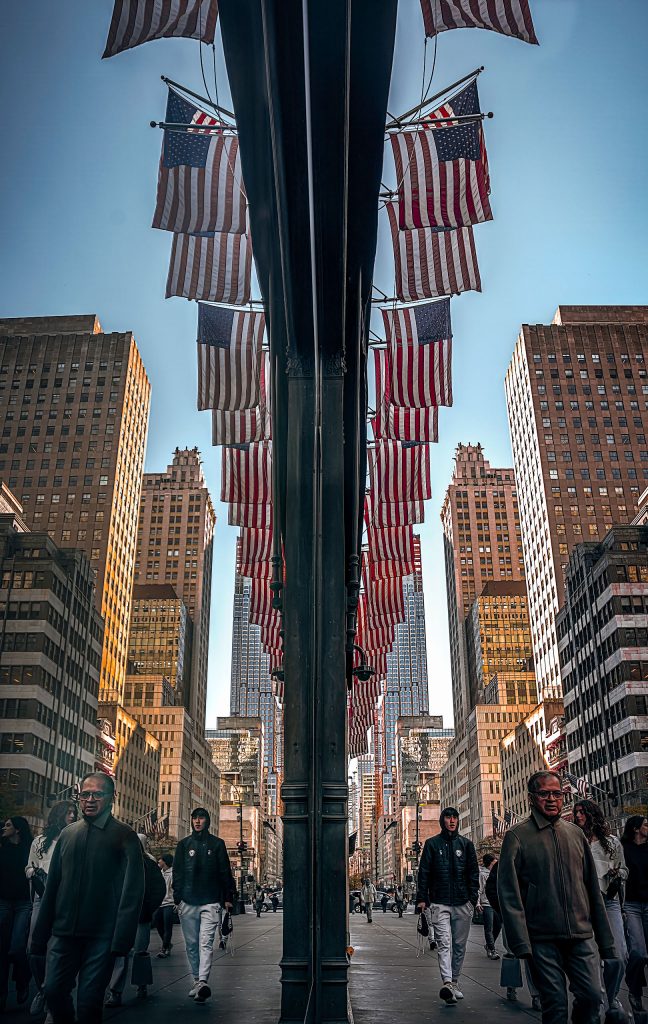 Komplexe, grafische Streetphotography auf der 5th Avenue in New York, basierend auf einer optischen Täuschung durch Spiegelung. Ein massiver, dunkler Balken – der Rahmen eines Schaufensters – zieht sich zentriert durch das Bild und teilt die Komposition. In der Glasscheibe spiegelt sich die belebte Straßenszene: Zahlreiche Passanten eilen vorbei, während sich im Hintergrund die typische, dichte Architektur der Wolkenkratzer erhebt. Mehrere amerikanische Flaggen ragen als markante Farbtupfer in das Spiegelbild. Das Zusammenspiel aus dem realen, dunklen Rahmen im Vordergrund und der detailreichen Spiegelung im Hintergrund erzeugt eine faszinierende Tiefe und visuelle Komplexität.