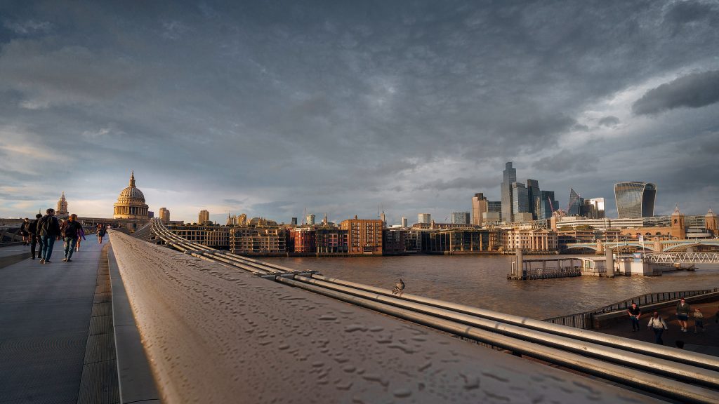 Fine-Art-Fotografie: Weitwinkelblick über die Millennium Bridge am Abend, belebt durch vereinzelte Fußgänger. Die stählerne Konstruktion führt den Blick diagonal zur links positionierten St. Paul’s Cathedral. Rechts ist das beleuchtete Themseufer mit Gebäuden zu sehen.