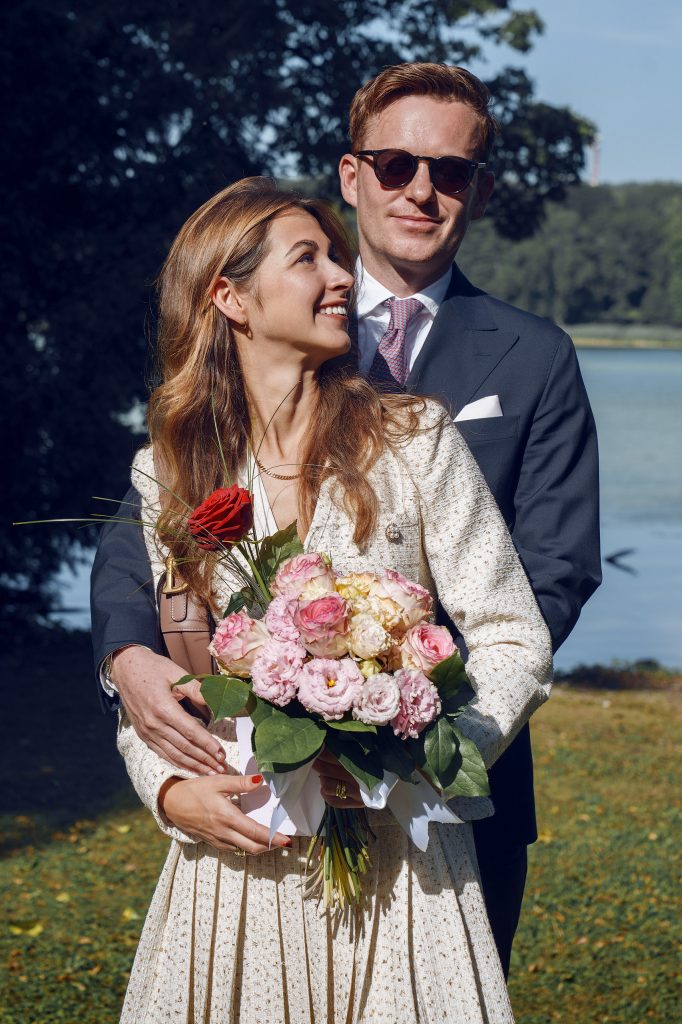 Emotionales Brautpaar-Portrait am Wasser: Eine Braut blickt mit einem roten Rosenstrauß in der Hand zärtlich zu ihrem Partner auf, der direkt hinter ihr steht, umrahmt von der idyllischen Kulisse eines Sees.