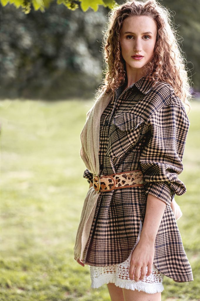 Mode-Shooting im Grünen: Modell mit langen Locken trägt eine sportliche karierte Holzfällerjacke mit auffälligem Taillengürtel und einen hellen Spitzenrock vor dichtem, grünem Hintergrund.