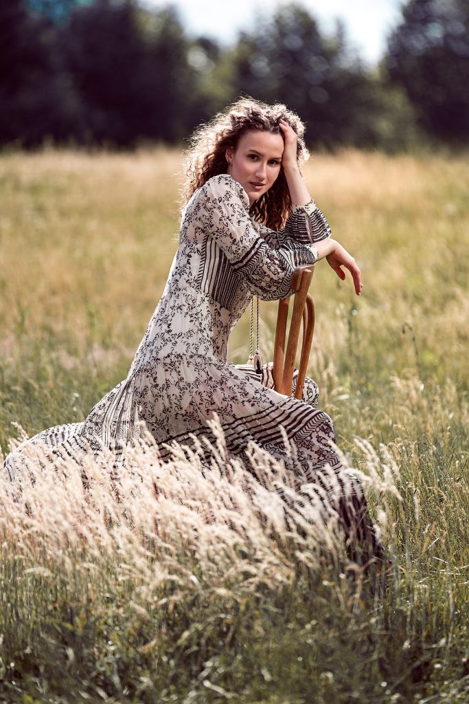 Romantisches Outdoor-Modefoto in warmem Licht. Ein Modell mit langen, weichen Locken sitzt entspannt auf einem einfachen Stuhl inmitten einer Wiese aus hohem, gelbem Gras. Sie trägt ein verspieltes, romantisches Kleid und hat die Arme lässig auf den Lehnen des Stuhls abgelegt. Die spätsommerliche Atmosphäre und ihre ruhige Pose strahlen eine friedliche, nostalgische Stimmung aus.