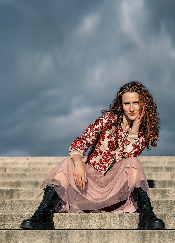 Ein starkes Mode-Porträt im Stilbruch vor einem strahlend blauen Himmel. Ein Modell mit langen, leuchtend roten Locken sitzt selbstbewusst auf den Stufen einer Steintreppe. Sie trägt ein extrem gegensätzliches Outfit, bestehend aus einer glamourös bestickten Jacke und einem voluminösen, hellen Tüllrock. Dazu trägt sie als derben Kontrast schwarze, klobige Lederboots. Ihr Blick ist intensiv und herausfordernd direkt in die Kamera gerichtet.