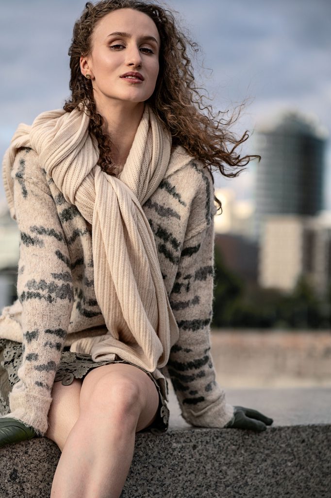 Urbanes Mode-Shooting: Modell mit langen Locken sitzt in einem kuscheligen Strickpullover und dickem Schal auf einer Mauer. Ein Spitzenrock ist bis zu den Oberschenkeln sichtbar. Der städtische Hintergrund ist verschwommen.