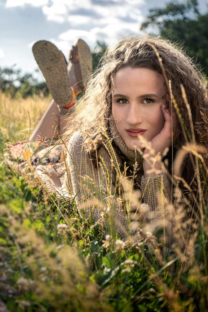 Stimmungsvolles Outdoor-Modefoto in warmer Atmosphäre. Ein Modell mit langen, weichen Locken liegt entspannt im hohen, gelblichen Gras einer herbstlichen Wiese. Sie trägt einen dicken, gemütlichen Rollkragenpullover, der sich farblich harmonisch in die herbstliche Umgebung einfügt. Der Hintergrund ist in einer weichen Unschärfe gehalten, wodurch der Fokus ganz auf der friedlichen Ausstrahlung des Modells liegt.
