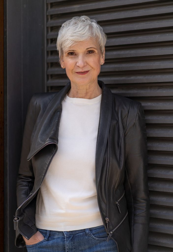 Charakterstarkes Fine-Art-Porträt einer älteren, modern gestylten Frau mit kurzen Haaren. Sie blickt mit einem sympathischen, herzlichen Lächeln direkt in die Kamera. Ihr lässiges Outfit aus Jeans und einer coolen Lederjacke hebt sich elegant vor dem schlichten, dunklen Hintergrund ab.